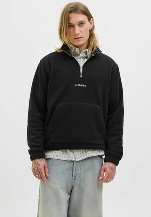 Sort fleece sweatshirt med lynlås krave, frontlomme og "JJ Studios" logo. Båret over en ternet skjorte. Casual design, blød tekstur.