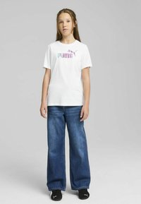 Camiseta blanca de algodón con logo "PUMA" multicolor, combinada con unos vaqueros de denim azul de pierna ancha y zapatillas deportivas negras.