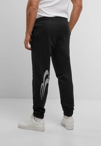 Black joggers with an elastic waistband, featuring a grey graphic design on the left leg. Paired with white sneakers for contrast.