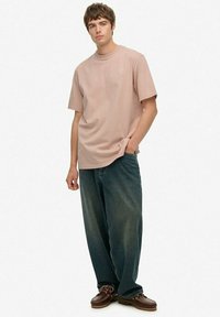 En ung mand står op, iført en løs lyserød T-shirt, vide mørke jeans og brune lædersko, med den ene hånd i lommen.