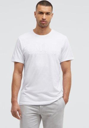 Man standing wearing a white CMP Urban Essentials T-shirt and light gray pants with one hand in pocket against plain background.