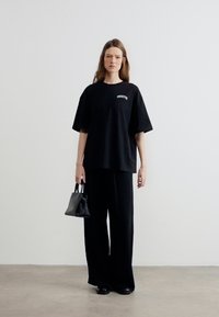 Γυναίκα που στέκεται μπροστά σε λευκό τοίχο, φορώντας χαλαρό μαύρο μπλουζάκι, φαρδιά μαύρα παντελόνια, κρατώντας μικρή μαύρη τσάντα στο δεξί χέρι.