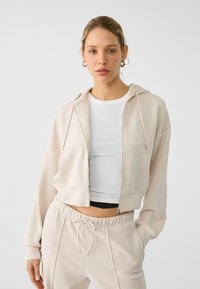 Skraćena zip-up dukserica od meke bež tkanine, s prednjim zatvaračem, kapuljačom i opuštenim krojem, u kombinaciji s odgovarajućim širokim hlačama.