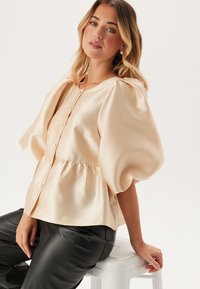 Blusa a maniche puff in morbida satin beige, con una chiusura frontale a bottoni e una vita arricciata, abbinata a pantaloni in pelle nera.