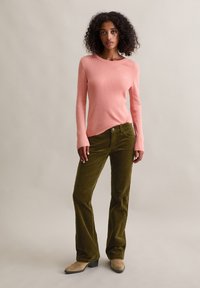 Frau mit lockigem Haar, die ein pinkfarbenes, geripptes Langarmshirt, olivgrüne Cordhosen und beige Stiefeletten trägt und vor einem einfachen Hintergrund steht.