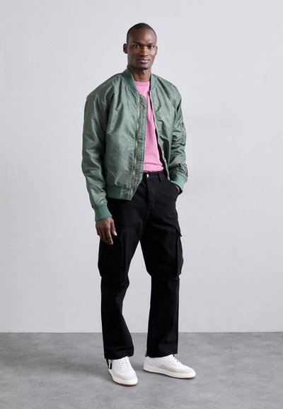 Veste bomber verte avec une texture brillante, t-shirt rose, pantalon cargo noir et baskets blanches avec des accents gris. Détail logo subtil sur la veste.