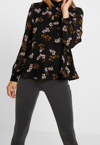 Schwarze Bluse mit langen Ärmeln, die mit pinken und braunen Blumenmustern verziert ist, mit Rüschensaum an Kragen und Ärmeln sowie einem Peplum-Taille-Design.