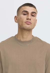 Brauner gerippter Sweatshirt mit Rundhalsausschnitt; getragen mit einer silbernen Kettenhalskette. Der Stoff erscheint weich und hat eine glatte Textur.