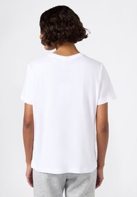 Persona con capelli ricci corti che indossa una maglietta bianca a maniche corte e pantaloni grigi chiari, di spalle su uno sfondo bianco.