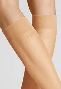 Hautfarbene Overknee-Strümpfe mit einer glatten, matten Oberfläche. Sie verfügen über ein breites, strukturiertes Band oben, auf dem "HUDSON" in heller Farbe gedruckt ist.