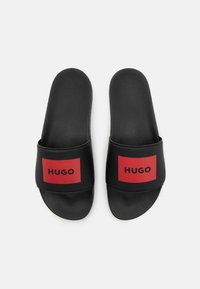 Svarte sklisikre sandaler med teksturert såle, med en bred svart stropp dekorert med en rød rektangulær panel som viser "HUGO" i svarte bokstaver.