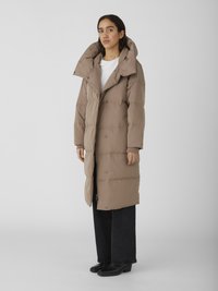 Manteau rembourré beige avec un col haut, présentant une fermeture éclair sur le devant et de grandes poches. Texture douce avec un design matelassé et un rembourrage isolant.