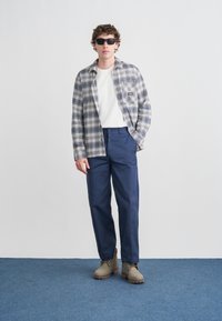 Καρό πουκάμισο σε λευκό, μπλε και γκρι χρώμα· λευκό T-shirt από κάτω· σκούρο μπλε παντελόνι· γκρι μποτάκια από σουέτ· σκούρα γυαλιά ηλίου· στέκεται μπροστά σε μονόχρωμο φόντο.