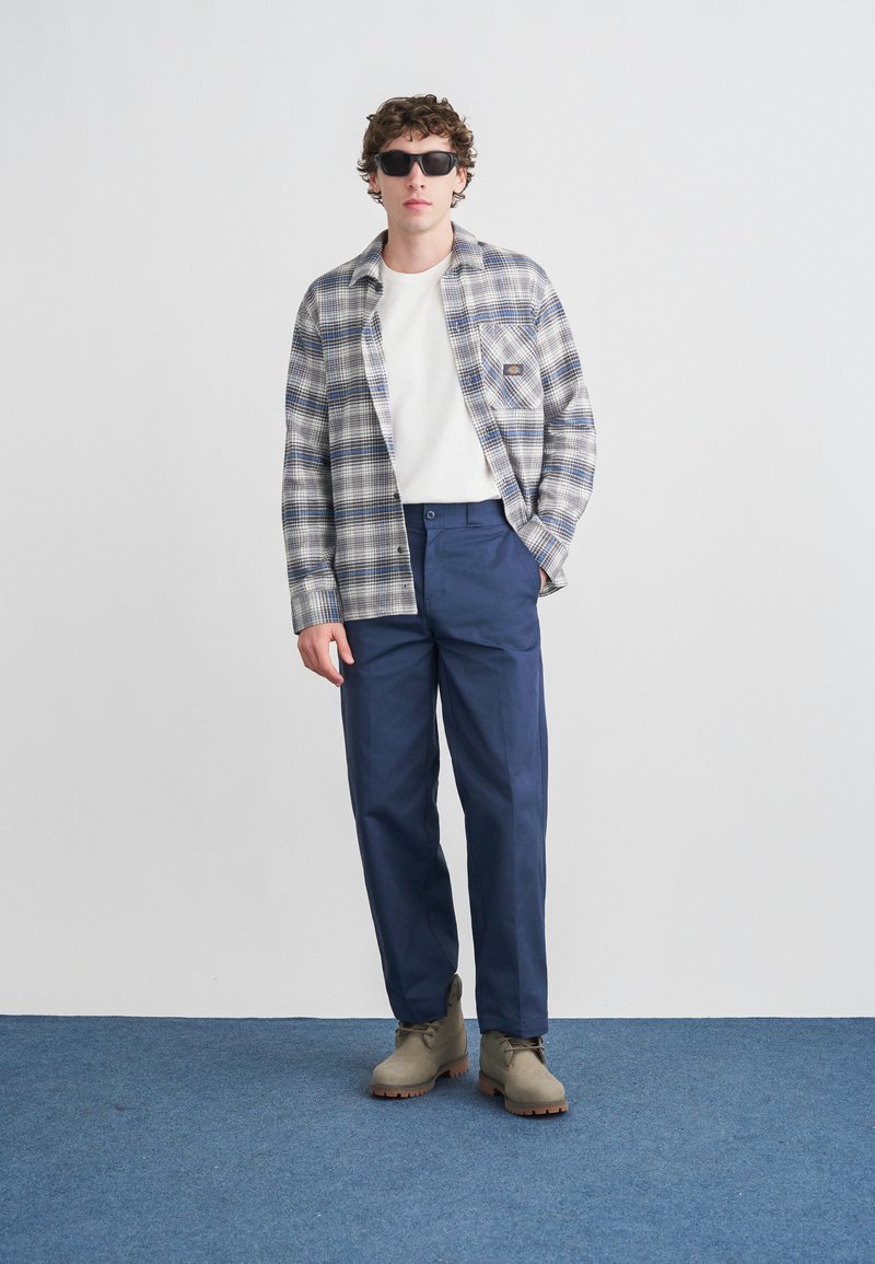 Kariertes Hemd in Weiß, Blau und Grau; weißes T-Shirt darunter; marineblaue Hose; graue Wildlederstiefel; dunkle Sonnenbrille; steht vor einfachem Hintergrund.