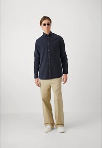 Navy-blaues Hemd mit langen Ärmeln, kombiniert mit beigen Hosen und weißen Sneakern. Das Model trägt Sonnenbrillen und steht vor einem einfarbigen Hintergrund.