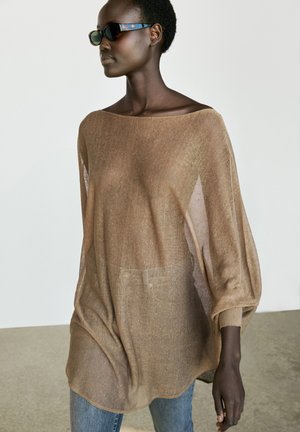 Femme portant un haut marron ample et transparent à manches longues, un jean bleu et des lunettes de soleil foncées devant un fond clair uni.
