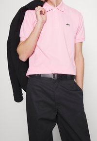 Camisa polo de color rosa claro con cuello clásico y dos botones, combinada con pantalones negros y una chaqueta negra colgada sobre un hombro.