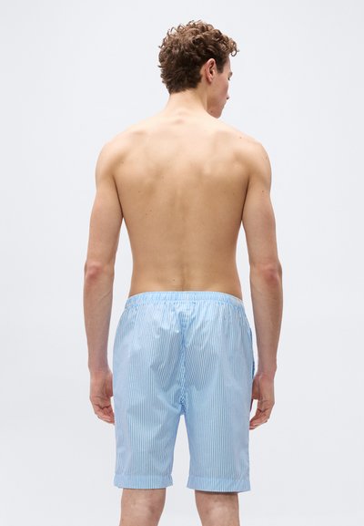 Νεαρός άνδρας με πλάτη φοράει σορτς με ανοιχτό γαλάζιο και λευκές ρίγες, σε απλό φωτεινό φόντο.