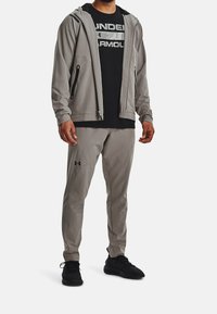 Grå sportjacka med dragkedjefickor, kombinerad med en svart Under Armour t-shirt med logotyp och grå byxor, kompletterad med svarta sneakers.