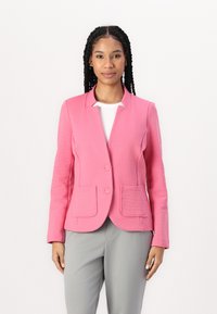 Růžový blazer se strukturovaným designem, dvěma předními kapsami a zapínáním na jedno knoflík, spárovaný se šedými kalhotami. Texturovaný povrch.