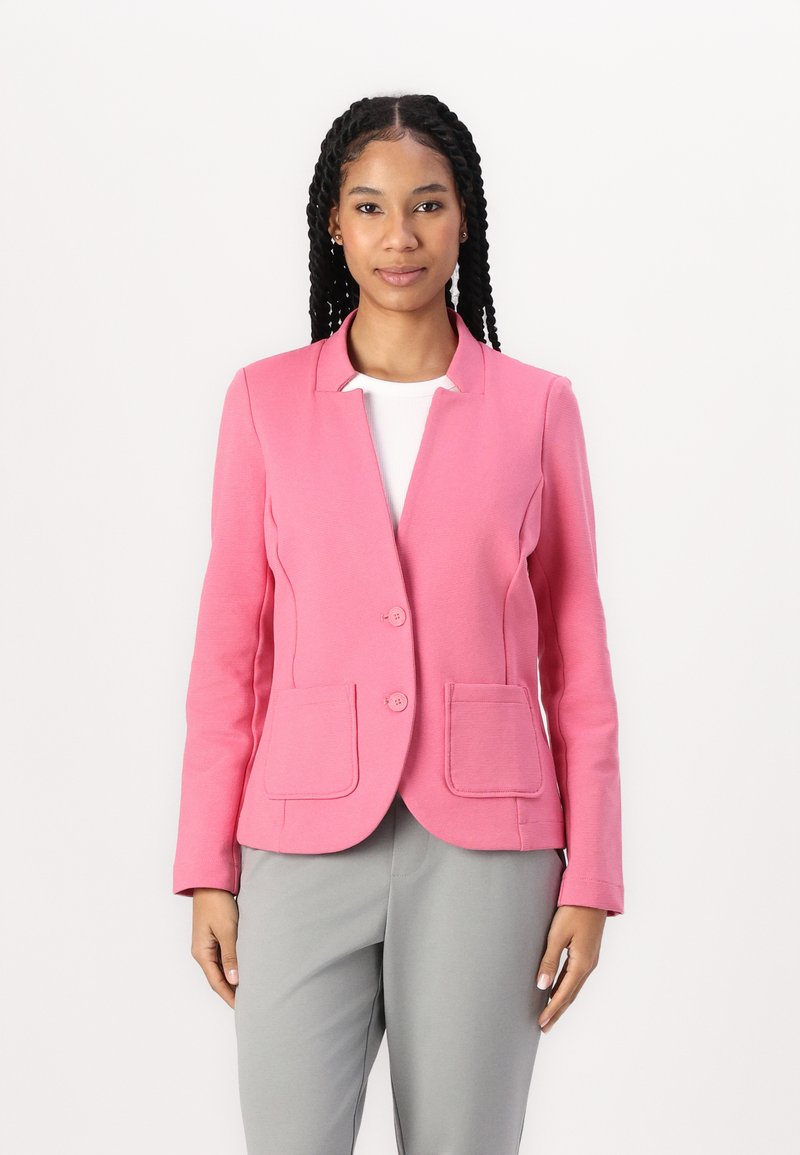Růžový blazer se strukturovaným designem, dvěma předními kapsami a zapínáním na jedno knoflík, spárovaný se šedými kalhotami. Texturovaný povrch.