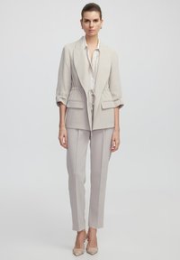 Beige taillierter Blazer mit tailliertem Schnitt, knöpfbaren Ärmeln und zwei Vorderreißverschlusstaschen, kombiniert mit passenden Slim-Fit-Hosen und beigen High Heels.