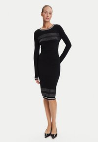 Abito in maglia nero con maniche lunghe, scollatura rotonda e dettagli a striscia argentata sul petto e sull'orlo. La silhouette aderente esalta la forma.