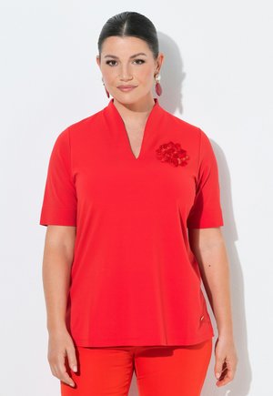 Frau mit dunklen Haaren in einem roten V-Ausschnitt-Top mit Blumenstickerei und passenden roten Hosen, die Perlen- und rote Tropfenohrringe trägt.