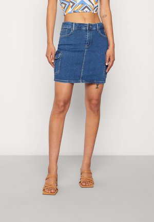 Femme portant une mini-jupe en jean bleu avec des poches latérales, un crop top géométrique multicolore, et des sandales à brides camel, debout sur un sol gris.