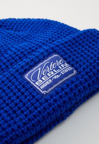 Blue knitted beanie featuring a textured pattern and a rectangular label with white text reading "Vertere," "BERLIN," and "HIGHER STATES."
