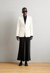 Weißer maßgeschneiderter Blazer über einem schwarzen Hemd mit Knöpfen, kombiniert mit einem langen schwarzen Rock. Die Accessoires umfassen dunkle Sonnenbrillen und schwarze Schuhe.