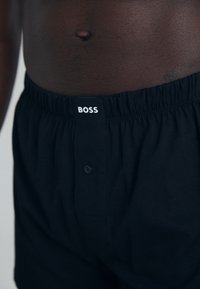 Čierne bavlnené boxerky s elastickým pásom, so štítkom s bielym logom "BOSS" a jedným gombíkom na prednej strane.