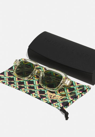 Gafas de sol transparentes con un marco verde translúcido y lentes oscuros, colocadas sobre una funda negra con un patrón colorido, al lado de un estuche negro.