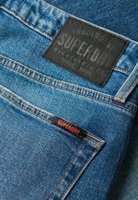Jeans de denim en azul medio con una etiqueta de cuero negro texturizado que dice "SUPERDRY" y una etiqueta de acento roja, con costuras en naranja.