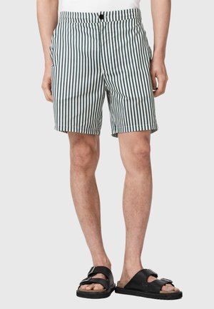 Mand iført hvide og mørke vertikalt striped shorts og sorte remsandaler, stående mod en ensfarvet lys baggrund.