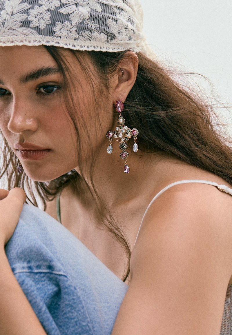 Jeune femme aux longs cheveux porte un foulard en dentelle blanche, des boucles d'oreilles pendantes colorées, un top blanc à bretelles et tient son genou enveloppé dans du denim bleu.