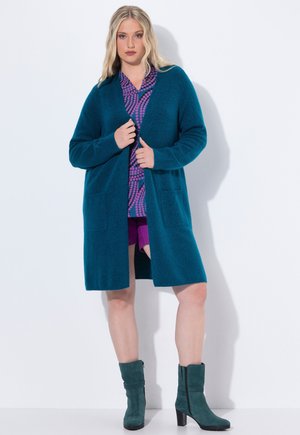 Cardigan teal con maniche lunghe, apertura frontale, tessuto testurizzato e due tasche frontali, indossato sopra un top viola a fantasia e pantaloni corti.
