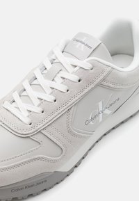Lys grå sneakers med en blanding av glatt skinn og semsket skinn, med hvite snørebånd og en sølvfarget logo aksent på siden. Teksturert gummisåle.
