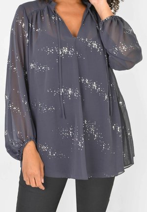 Vrouw draagt een losse, donkere semi-transparante blouse met zilveren pailletten sterrenpatronen en lange geplooide mouwen, gecombineerd met een zwarte broek.