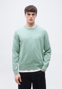Jeune homme aux cheveux bouclés portant un pull vert menthe par-dessus une chemise blanche et un jean foncé, debout devant un fond clair uni.