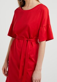 Vestido de algodón rojo con mangas cortas acampanadas, un escote redondo y una cinturilla con cinta. Textura suave, diseño simple, sin patrones notables.