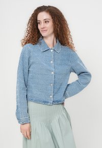 Μπλε cropped μπουφάν με κυματιστή υφή, μακριά μανίκια, κουμπιά μπροστά και γιακά, σετ με μια πλισέ φούστα ανοιχτού πράσινου χρώματος.