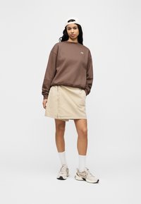 Καφέ φ ούτερ με λογότυπο, μπεζ τζιν φούστα, λευκές κάλτσες και ανοιχτόχρωμα αθλητικά παπούτσια. Διαθέτει χαλαρή εφαρμογή και απαλή υφή.