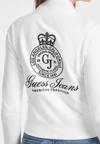 Bílá mikina s černou vyšívanou logem na zadní straně, s textem "Guess Jeans" a "American Tradition." S designem s zipovým límcem.
