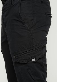 Närbild av svarta cargobyxor som visar framfickan, sidfickan med lock och knapp, samt detaljerad sömnad på tyget.