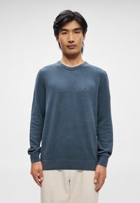 Blå, strikket genser med tekstur, rund hals og lange ermer, med subtil merkevare på brystet. Kombiner med lyse bukser.