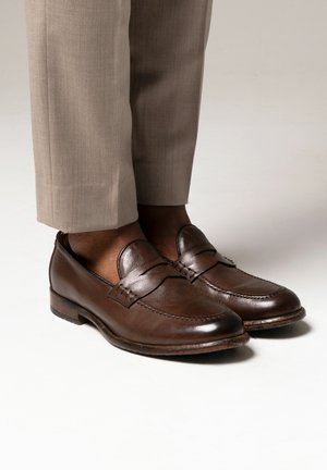 Mocasines de cuero marrón con punta redondeada, costuras decorativas y una textura suave. Tienen un tacón bajo y se combinan con pantalones beiges.