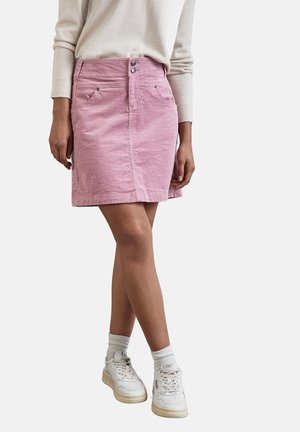Henkilö, jolla on pinkki korduroinen minihame, kermanvärinen neule, valkoiset nilkkasukat ja valkoiset lenkkarit, joissa on beige pohja.