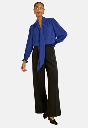 Royal blue blouse with a tied collar and puffed sleeves, paired with black wide-leg trousers and black pointed heels. Smooth fabric.