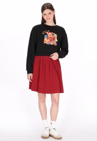 Fekete, rövid fazonú pulóver színes grafikával, piros, pliszírozott szoknyával párosítva. A modell fehér zoknit és hétköznapi cipőt visel.
